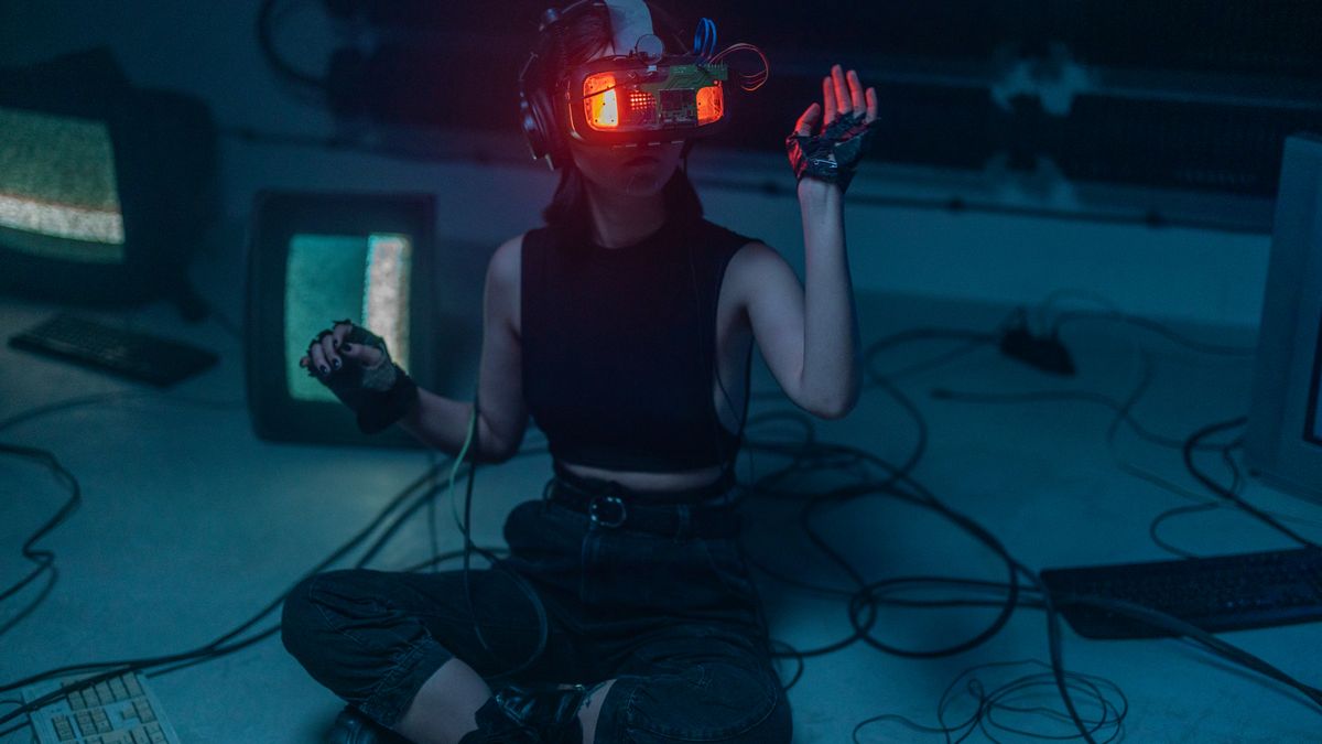 A woman wearing a VR headset sits on the floor surrounded by retro tech in a dark room.