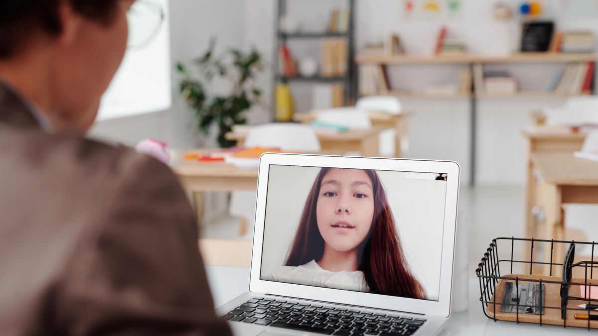 Teacher using laptop for virtual lesson with female student on screen in classroom setting.