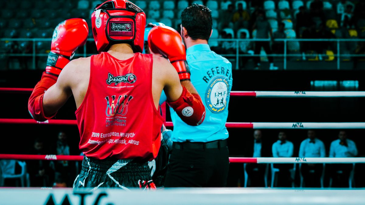 Rear view of a Muay Thai boxer in red gear facing the referee inside a boxing ring.
