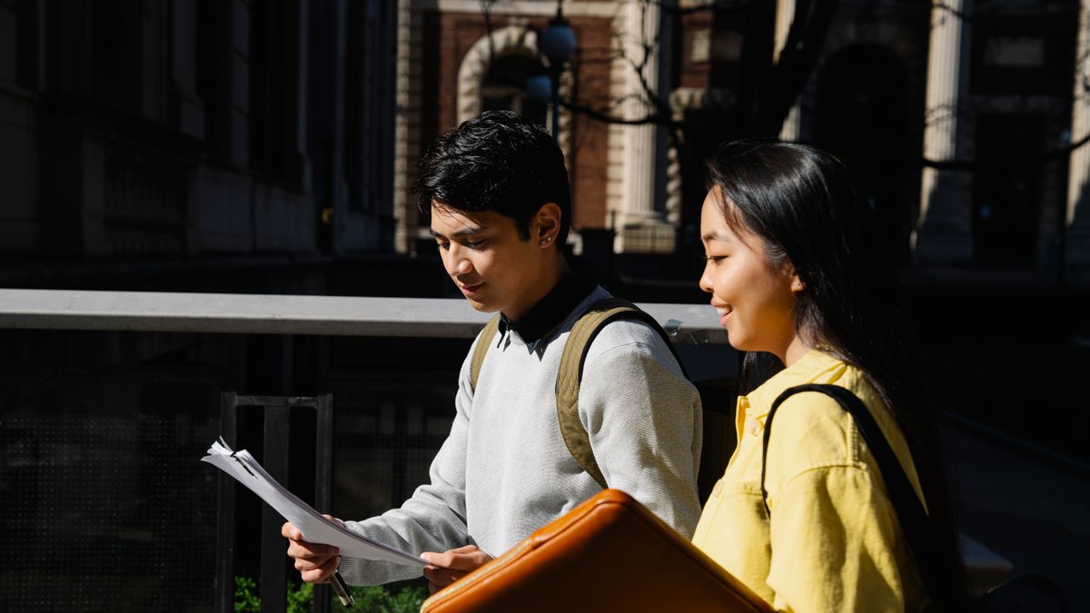 Two students walking with backpacks and papers on a bright day in the city.